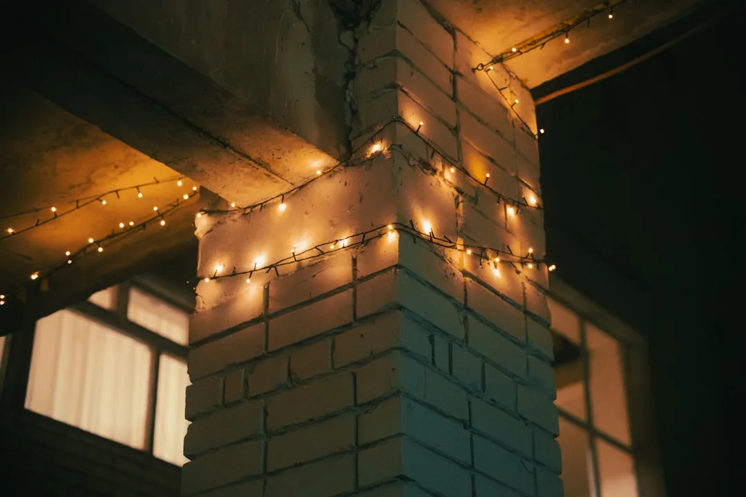 Warm string lights wrapped around a brick pillar at night, illuminating a building exterior with cozy golden glow