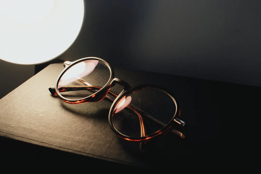Tortoiseshell eyeglasses resting on a book beside a glowing lamp, symbolizing low vision and reading light needs