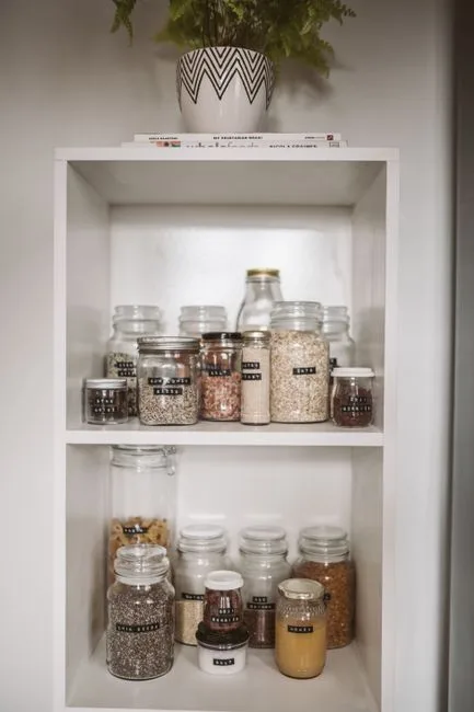 Organized white pantry shelves with labeled glass jars storing dry goods, grains, and spices in a tidy kitchen storage s