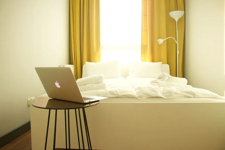 MacBook laptop on side table beside neatly made white bed in dual-purpose bedroom workspace transition setup