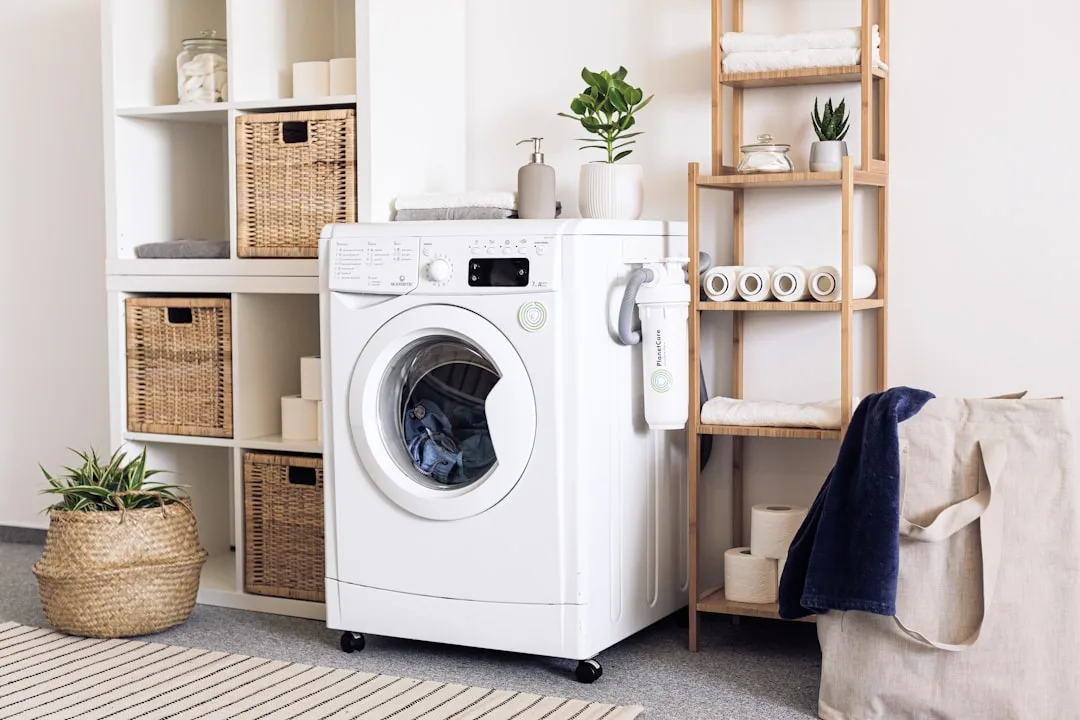 Compact laundry room with front-load washing machine, wooden shelving, wicker baskets, and microplastic filter device