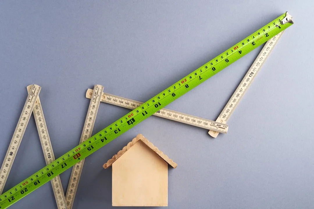 Green tape measure and folding ruler arranged as upward arrow above wooden house model on gray background