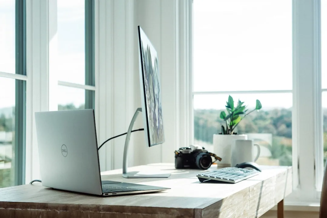 Dell laptop and monitor on a wooden desk positioned in front of large bright natural light windows for video calls