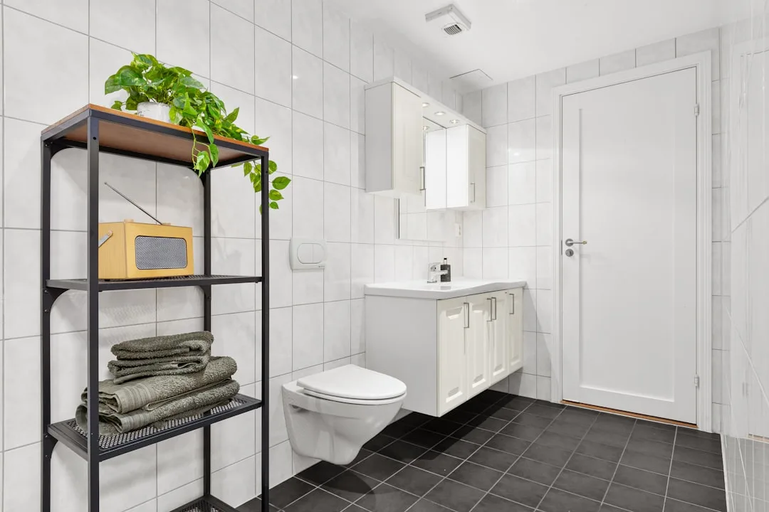 Modern white bathroom with wall-mounted toilet and black metal shelf unit storing towels and plant above toilet area