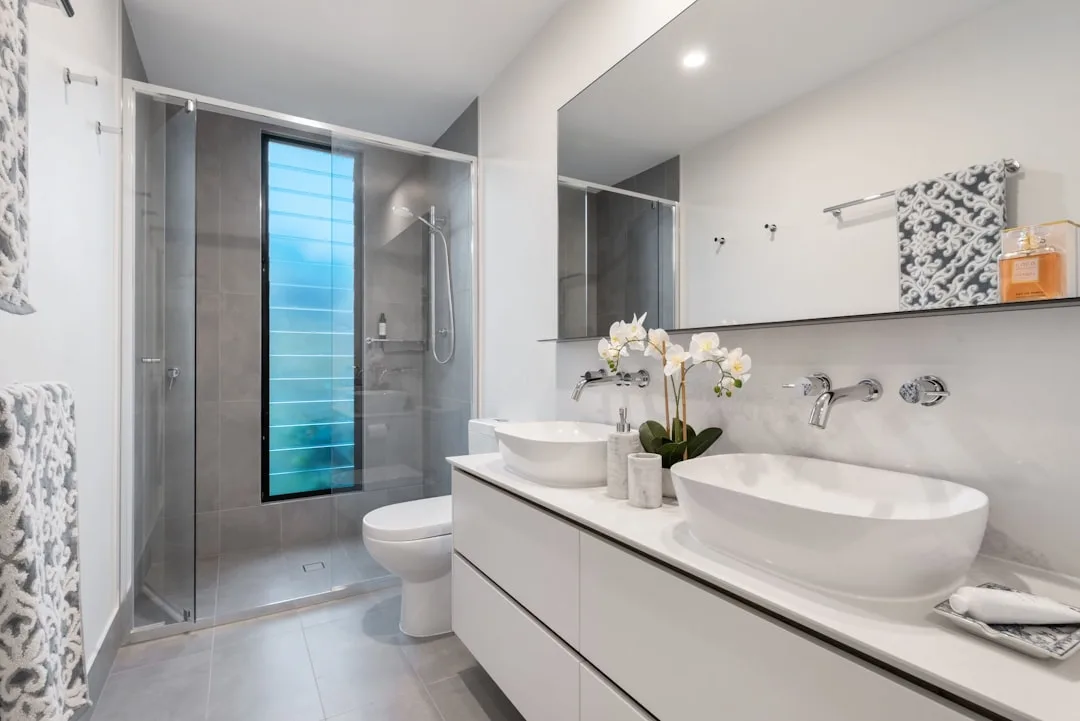 Modern tiled shower enclosure with glass door in contemporary white and grey bathroom with double vessel sinks