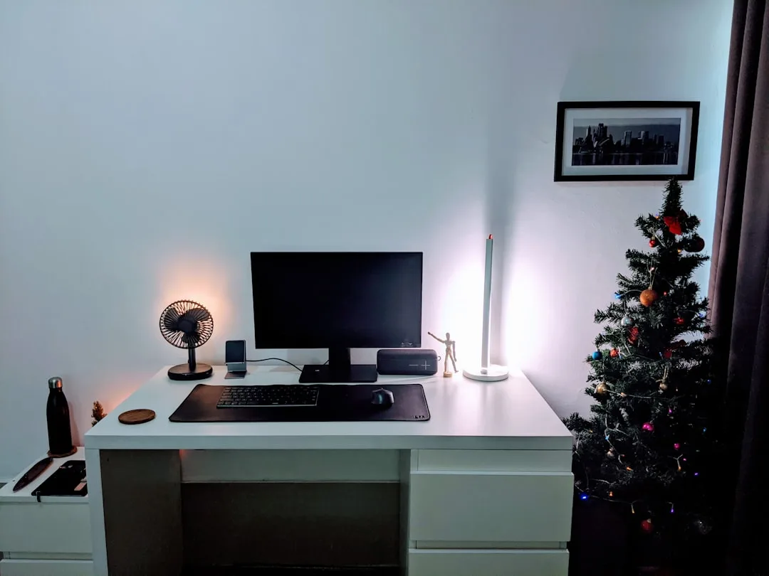 Minimalist white desk setup with monitor, keyboard, desk lamp and fan positioned against a white wall in a home office