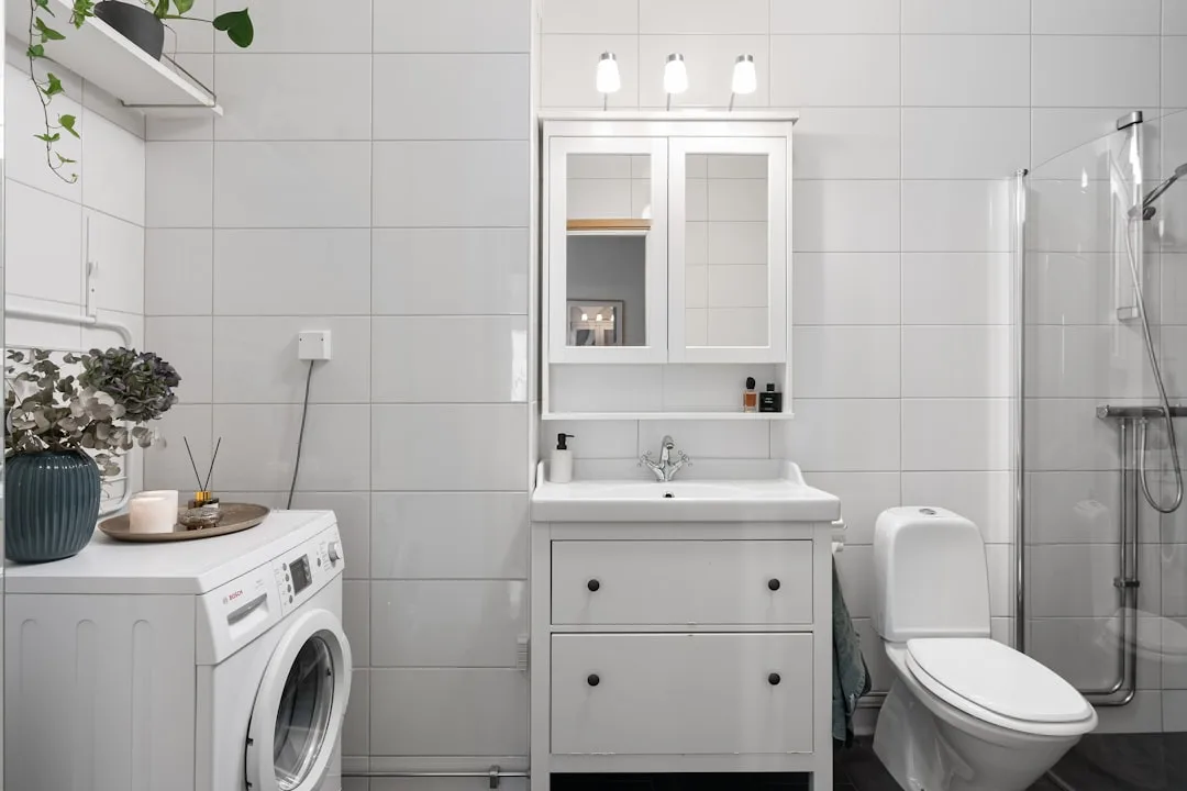 Small white bathroom with large tiles, vanity cabinet, washing machine and glass shower making space appear larger