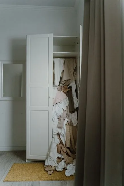 Open white wardrobe closet overflowing with piled beige and white clothing showing seasonal storage overflow problem