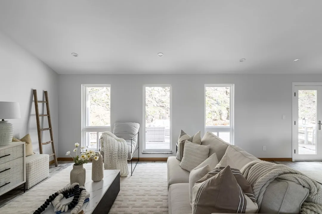 Bright living room with cool white walls, natural light from three windows, neutral sofa and warm wood accents