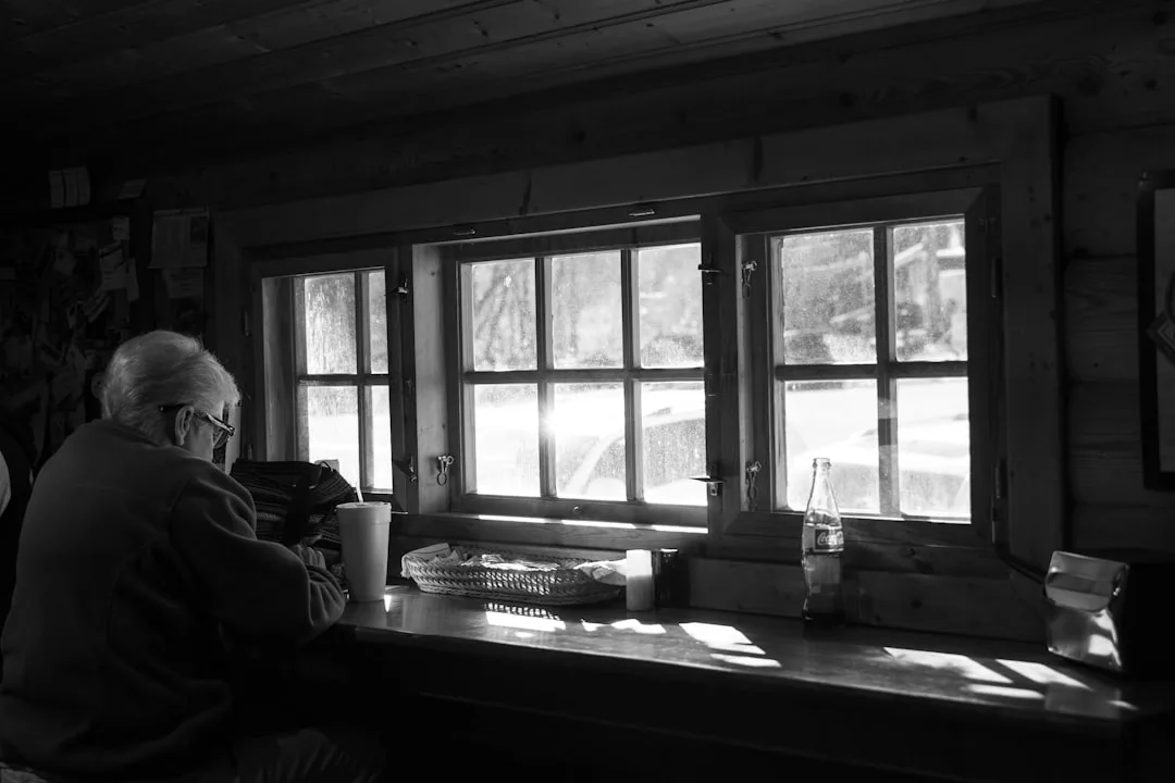 Elderly person sitting at a wooden window bench counter with colonial-style multi-pane windows in a rustic cabin interio