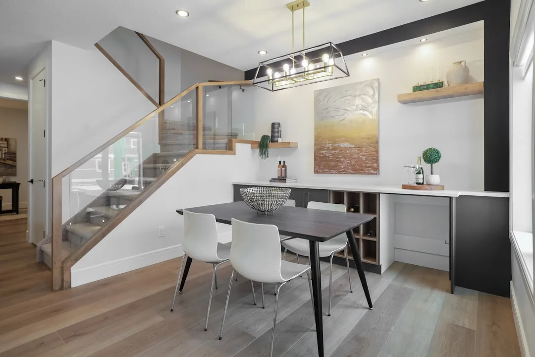 Modern open-plan dining area with dark table, white chairs, pendant light, and built-in shelving beside glass staircase