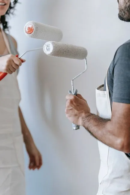 Two people in aprons holding paint rollers ready to paint a white wall in a north-facing room