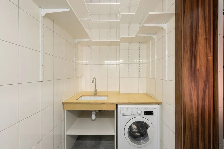 Compact laundry room with white wall-mounted shelving system, wooden countertop sink and under-counter washing machine