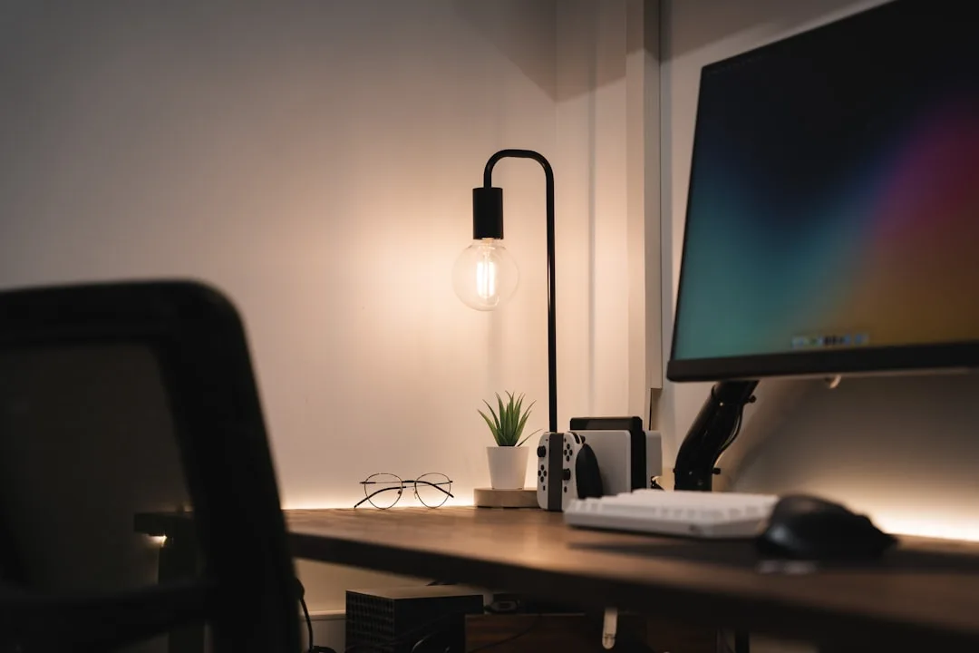 Modern home office desk with Edison bulb lamp, monitor, Nintendo Switch, and small plant in dim ambient lighting setup