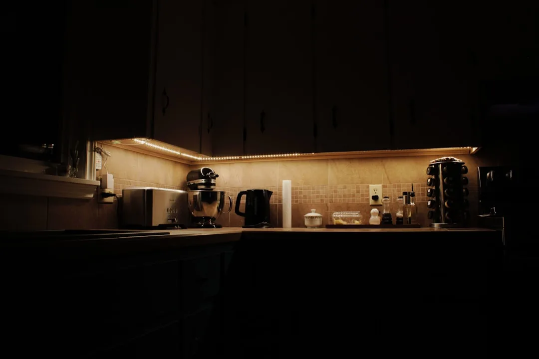 Under-cabinet LED strip lighting illuminating kitchen counter with stand mixer, kettle, and toaster in dark kitchen
