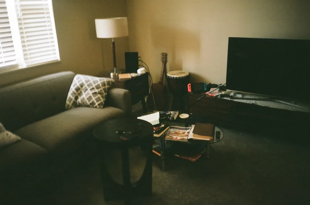 Living room with sofa pushed against wall, cluttered coffee table, TV stand, lamp and guitar showing common furniture pl