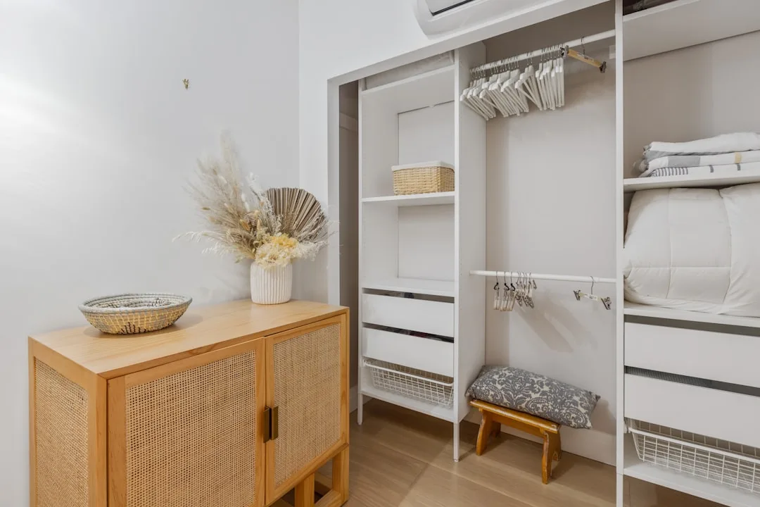 Organized white built-in closet with empty hangers, shelves, and drawers next to a rattan wood cabinet with dried flower