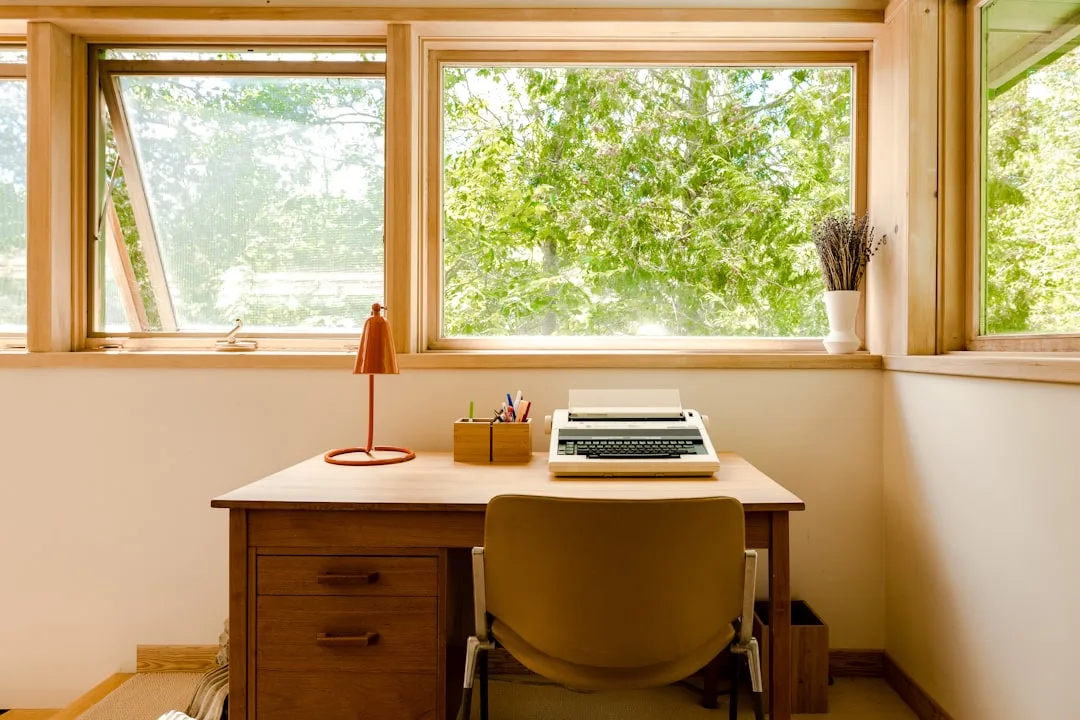 Wooden desk with typewriter positioned facing a large window with natural light and green trees outside
