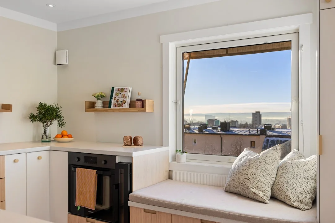 Built-in window seat bench with cushion and pillows beside kitchen oven, showing deep window casing and city view