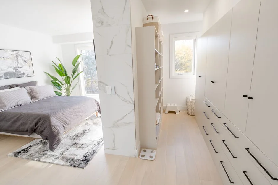 Walk-through wardrobe with built-in white cabinets and drawers connecting to a modern bedroom with marble accent wall