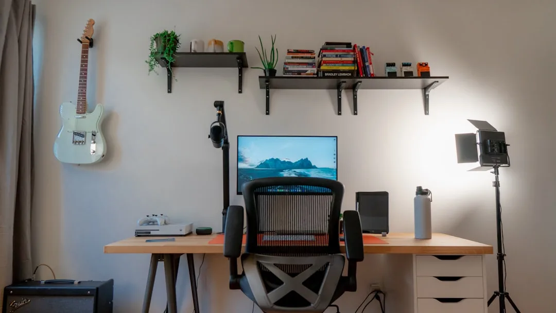 Compact home office desk setup with monitor, ergonomic chair, microphone arm, studio light, guitar, and wall shelves