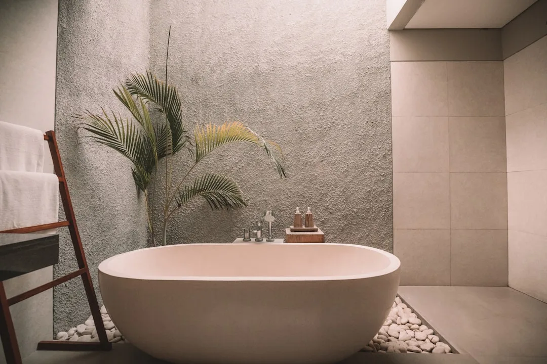 Freestanding oval soaking tub in luxury spa bathroom with stone pebble surround, palm plant, and towel ladder