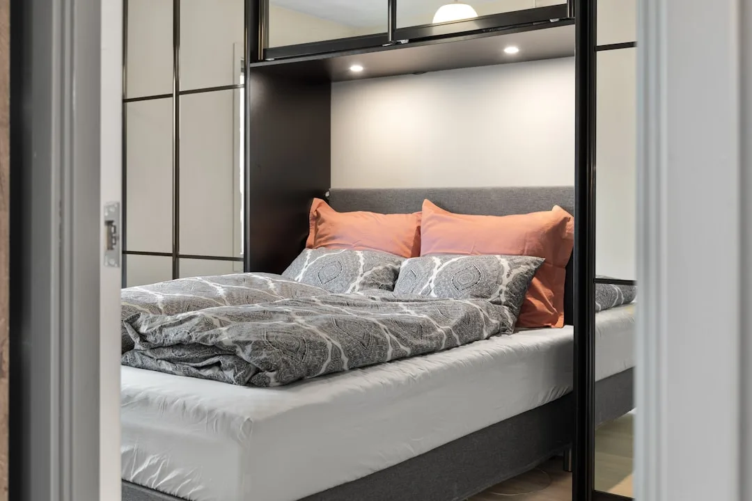 Murphy bed with gray upholstered headboard, orange pillows, and patterned duvet in a compact studio bedroom setup