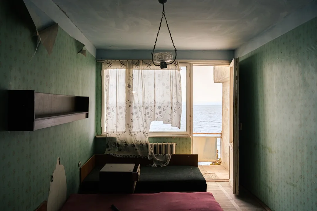Abandoned bedroom with mismatched worn furniture, green wallpaper, and ocean view window with sheer curtains
