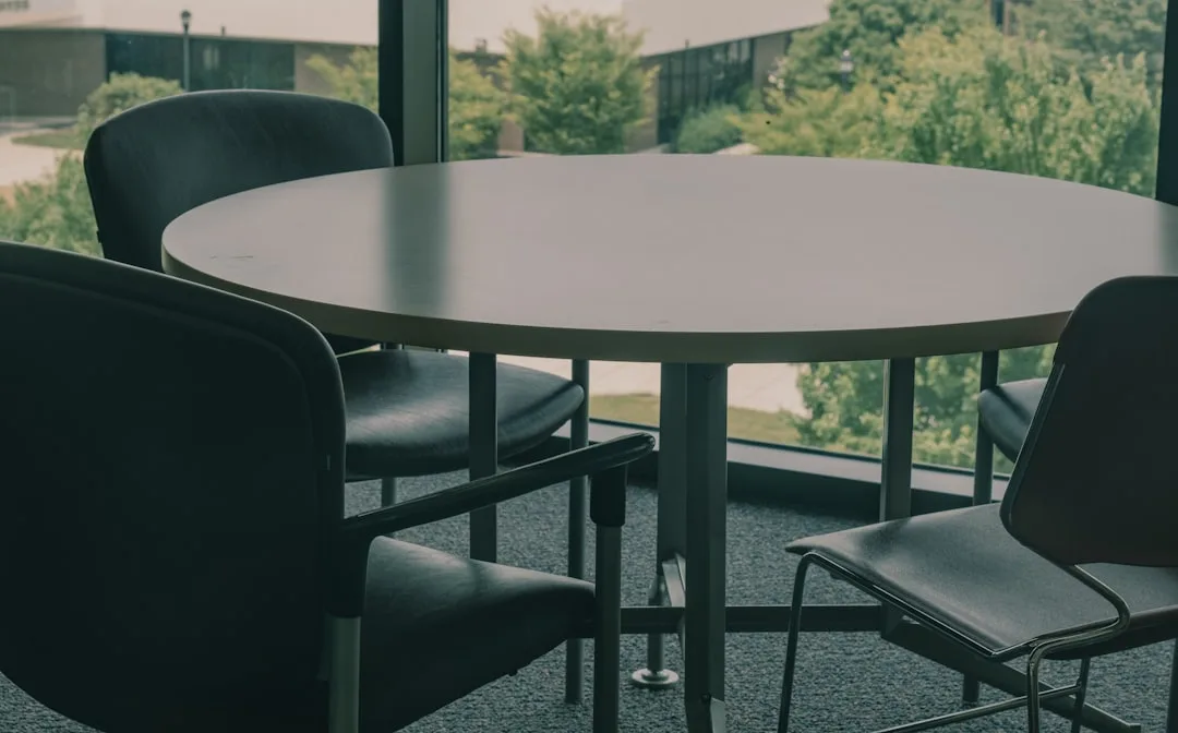 Round conference table with black chairs positioned near floor-to-ceiling windows in a small meeting room