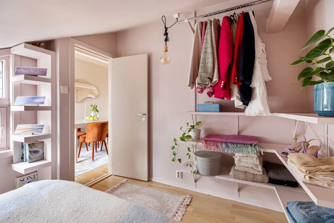 Open closet with hanging clothes rail, white shelves with folded blankets and storage boxes in a pink bedroom