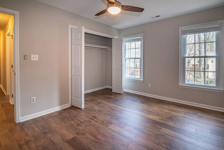 Empty bedroom with dark hardwood floors, open closet, ceiling fan, and two windows with natural light