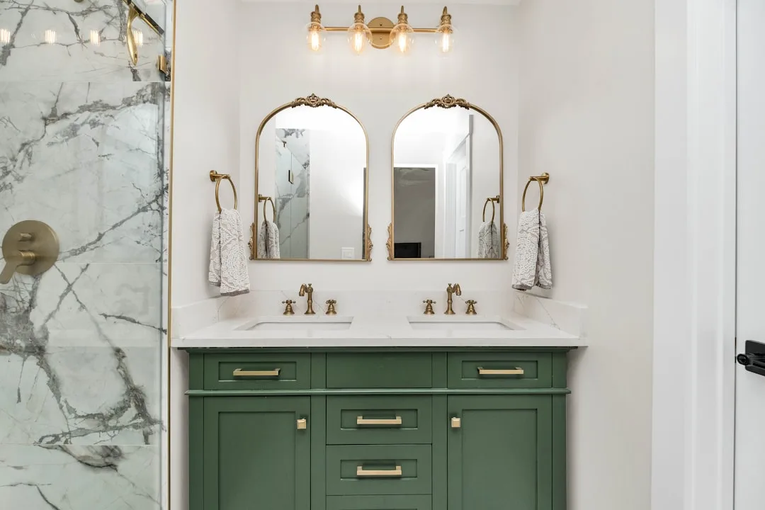 Elegant full bathroom with green double vanity, gold fixtures, marble shower, and arched mirrors showing renovation cost
