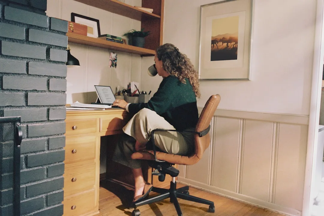 Woman working at wooden home office desk with styled shelf and laptop, seen from behind in cozy home workspace