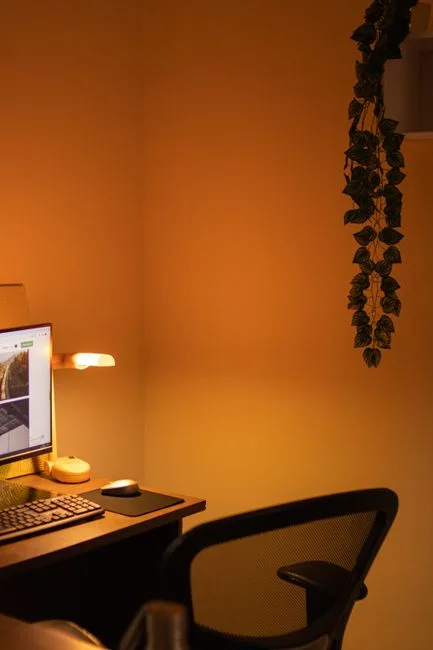 Home office desk setup with warm amber lighting, computer monitor, desk lamp, and hanging ivy plant against orange wall