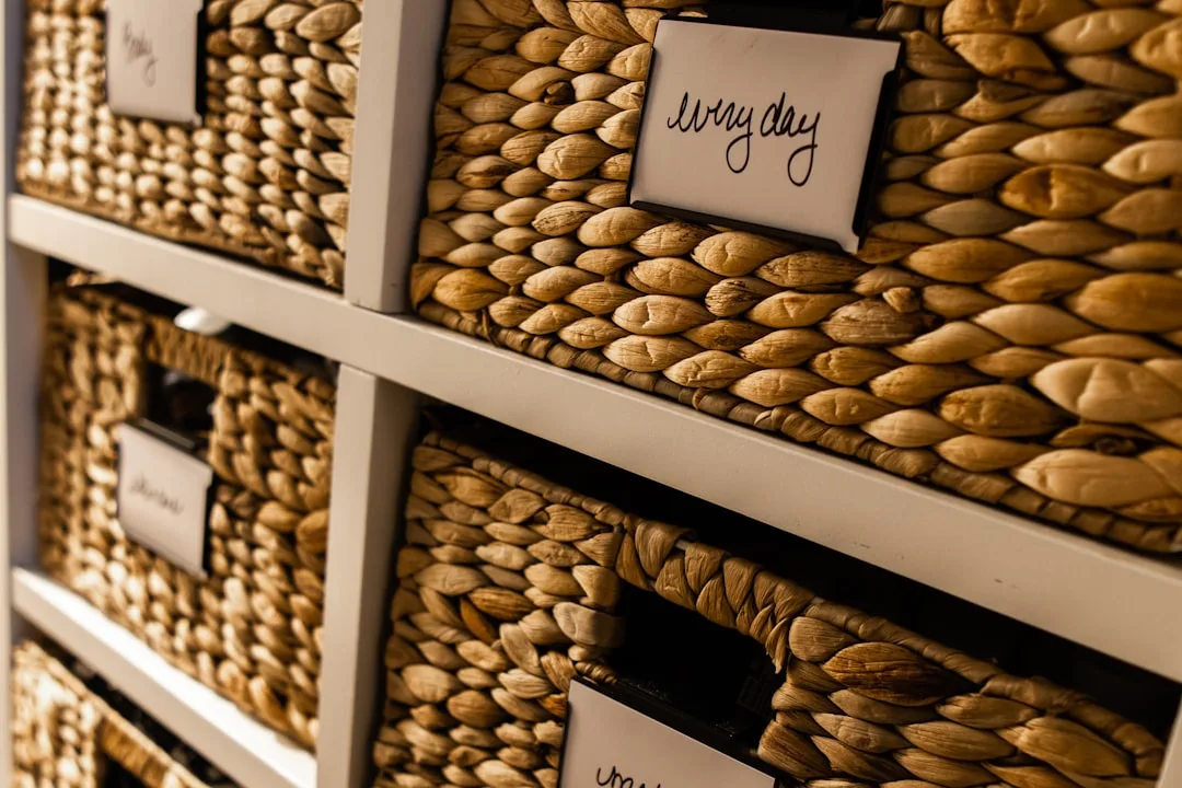 Woven basket storage bins organized on white shelves with handwritten labels for zone-based open shelf organization