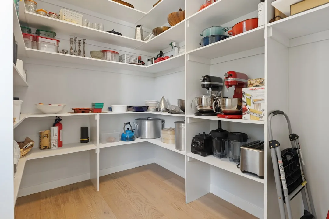 Organized walk-in pantry with white shelving storing kitchen appliances, cookware, and food storage containers