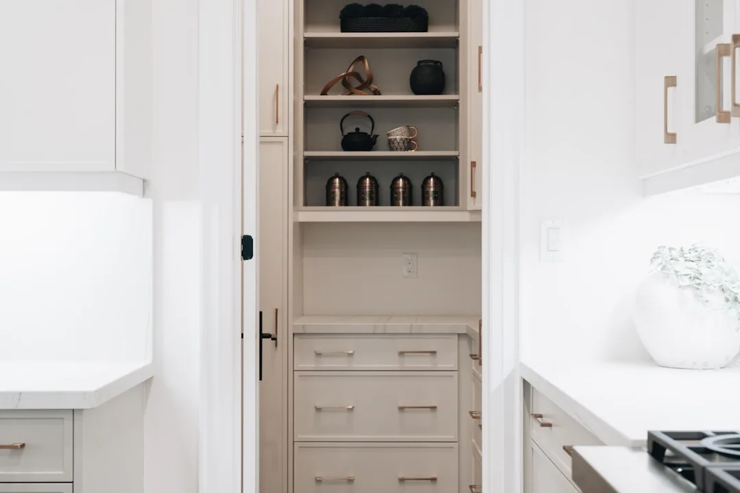 Open narrow pantry cabinet with vertical shelves displaying copper canisters, black teapot, and decorative items in whit