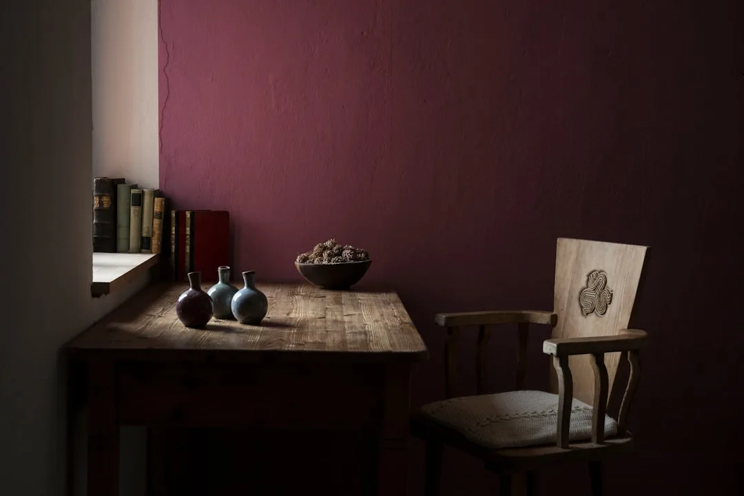 Deep burgundy wall in a rustic study with wooden desk, ceramic vases, and carved chair supporting creative work
