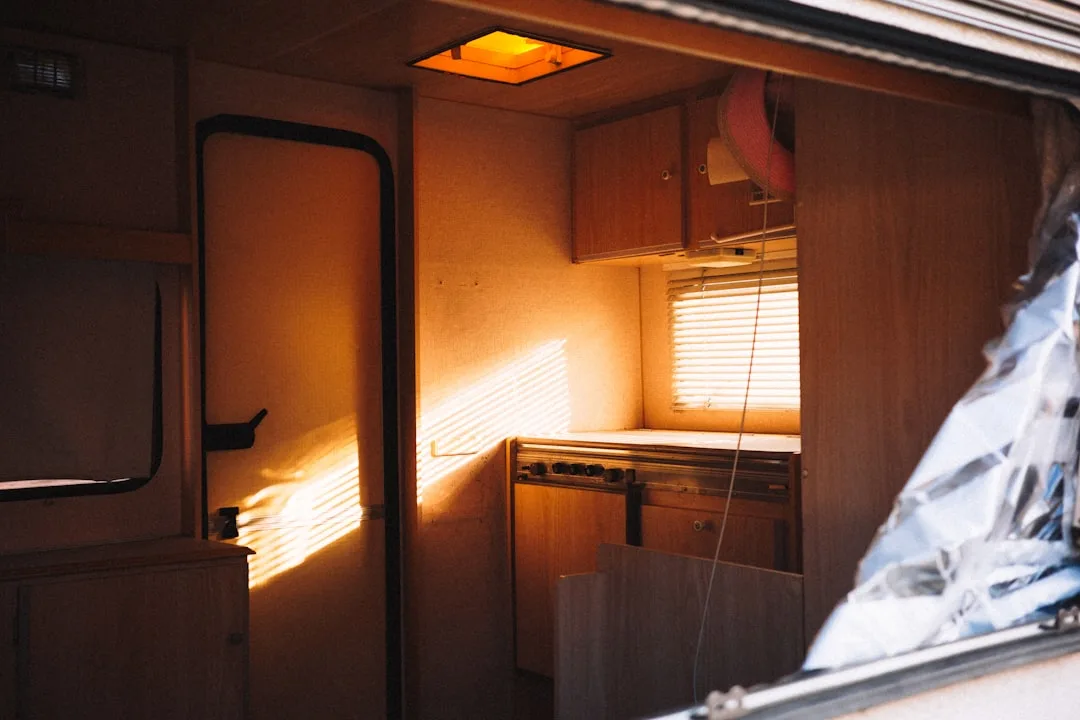 Warm sunlight streaming through blinds into a compact RV galley kitchen with wood cabinets and overhead vent lighting