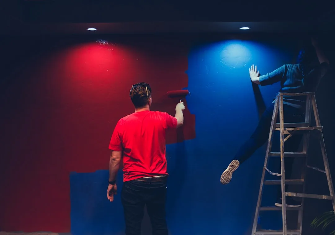 Two painters applying red and blue paint to walls using roller and hands, demonstrating color drenching technique indoor