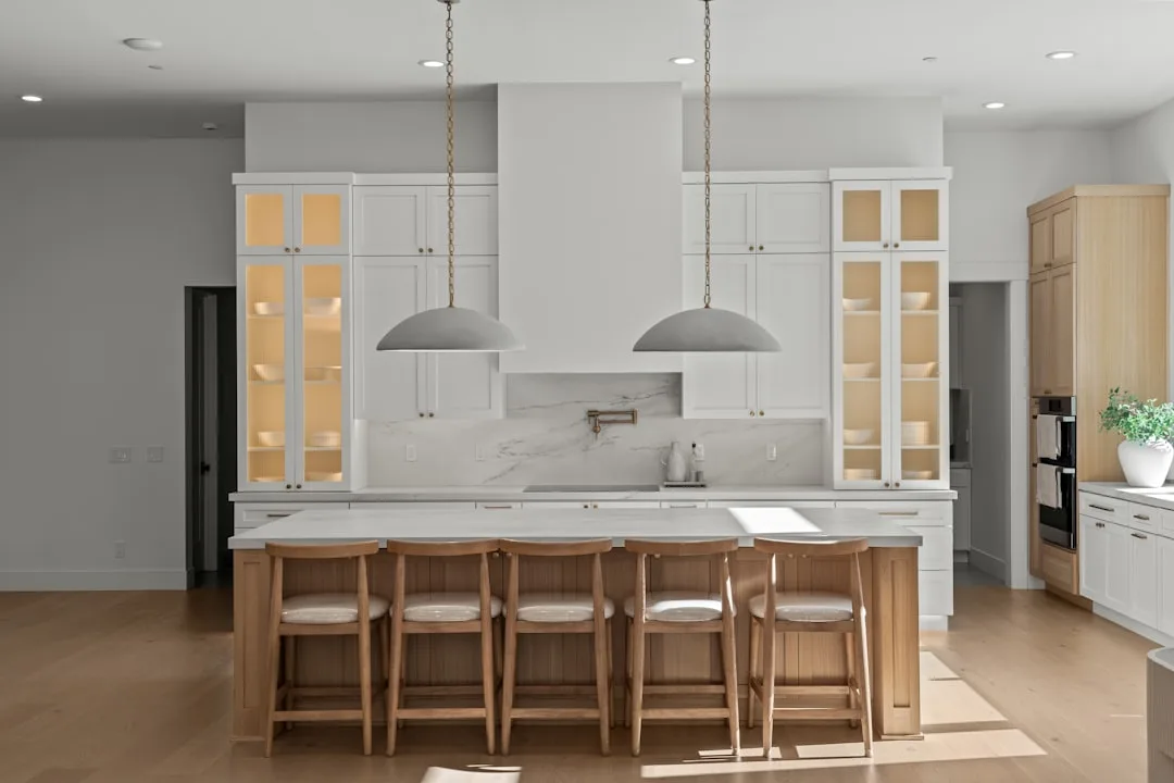 Modern white kitchen with large marble island, wood bar stools, pendant lights, and glass-front cabinets