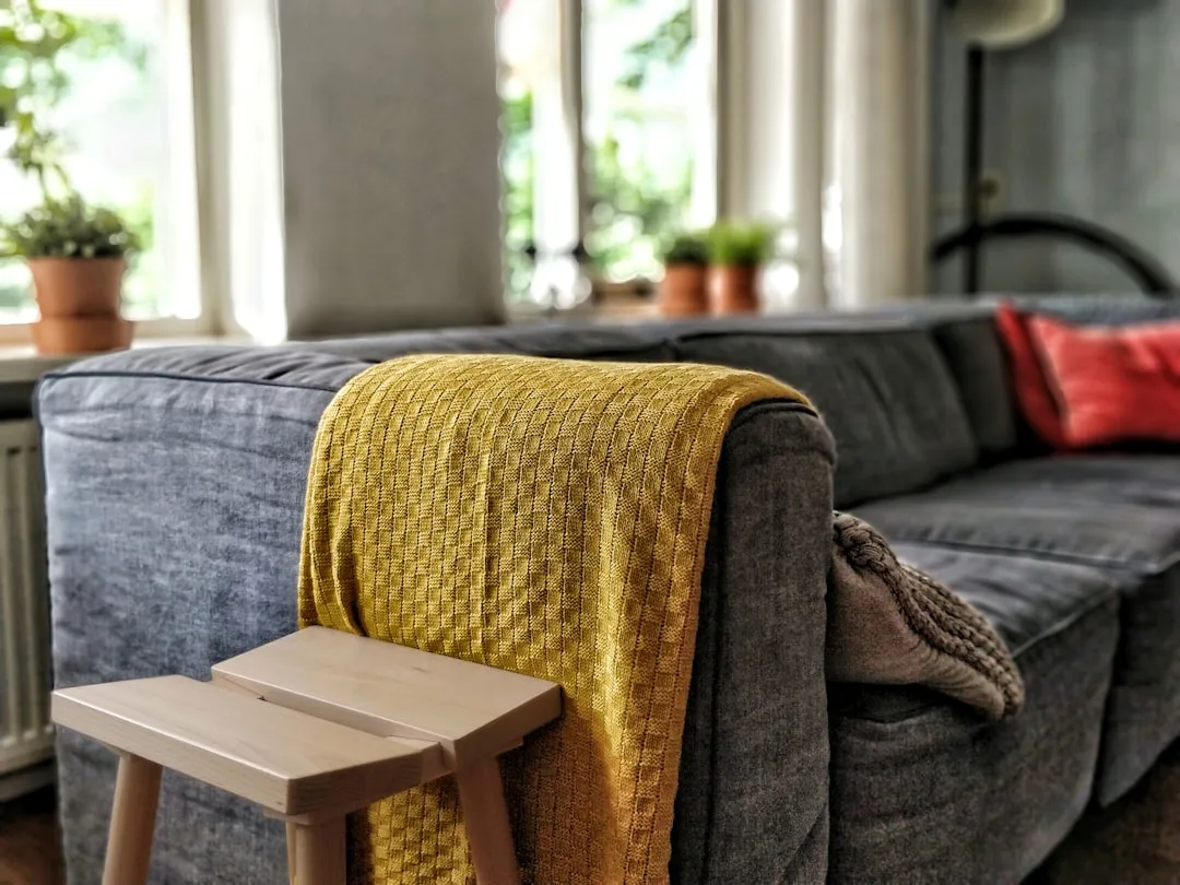 Yellow knit throw draped over gray sofa arm with red cushion and wooden stool in cozy living room