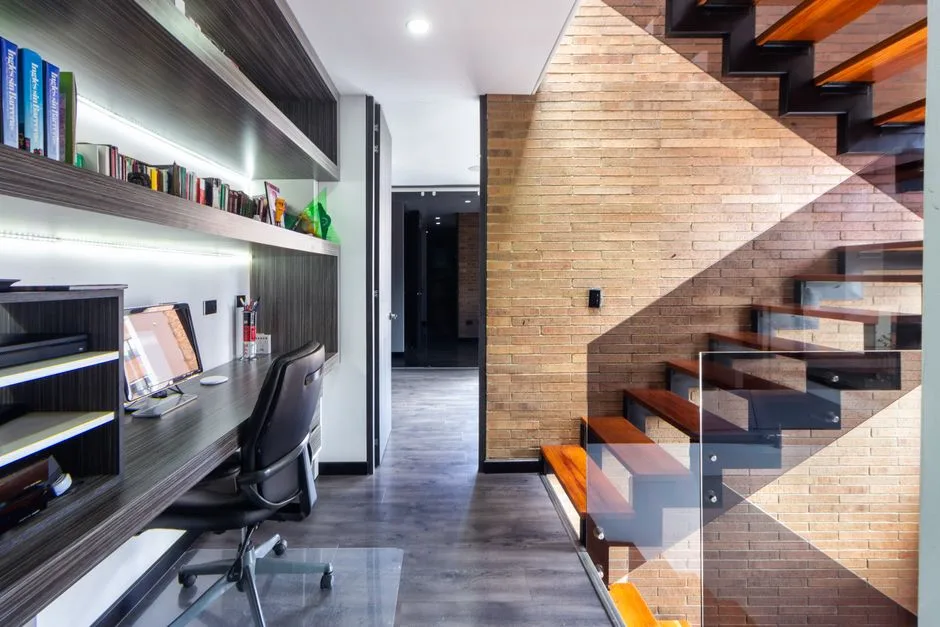 Functional under-stair home office with built-in shelving, desk, monitor, and ergonomic chair beside exposed brick wall