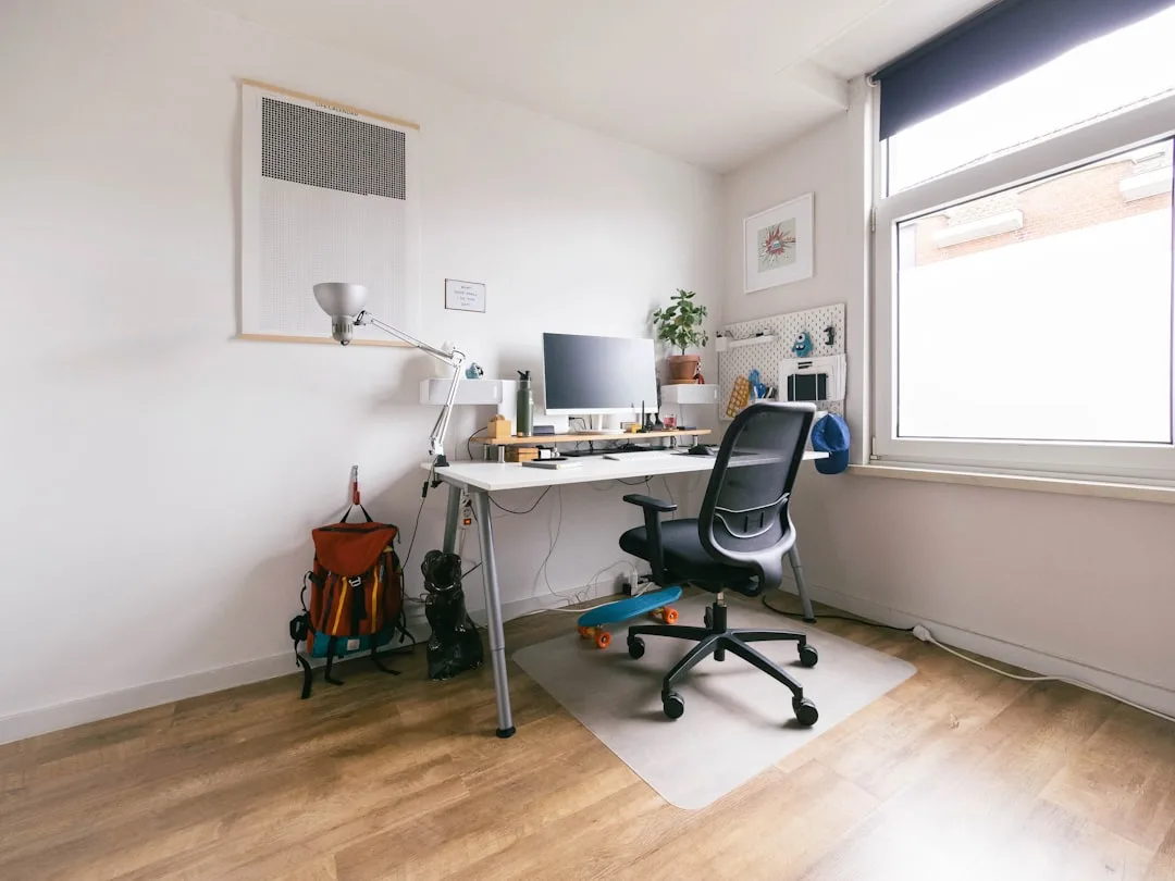 Black ergonomic mesh office chair at white desk in minimalist home office with wood floors and natural window light