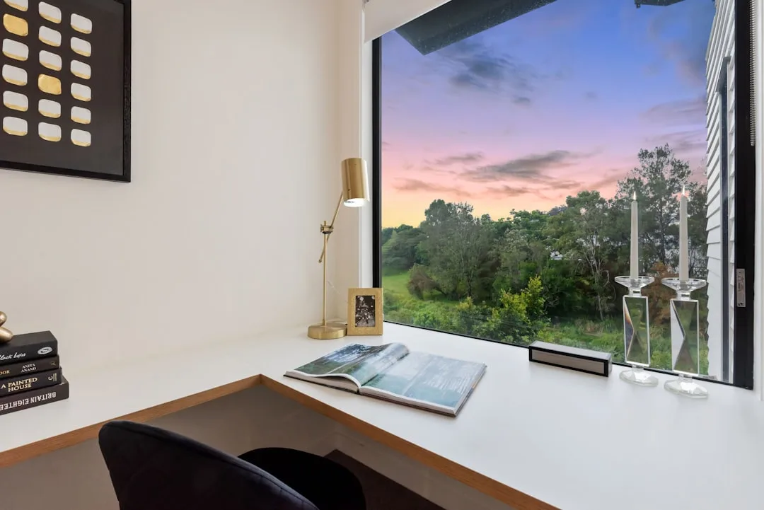 Modern built-in white desk nook with golden lamp, open book, and large window overlooking green trees at sunset