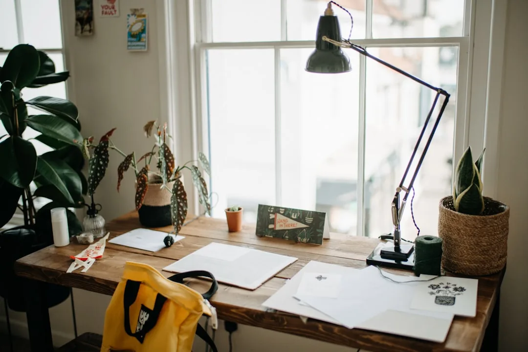 Rustic wooden home office desk by window with desk lamp, plants, papers and yellow bag in living room setup
