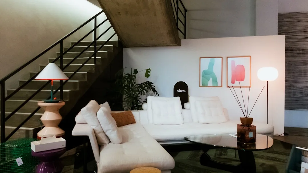 Modern living room with white sectional sofa, minimal decor, abstract wall art, and floor lamp under concrete staircase