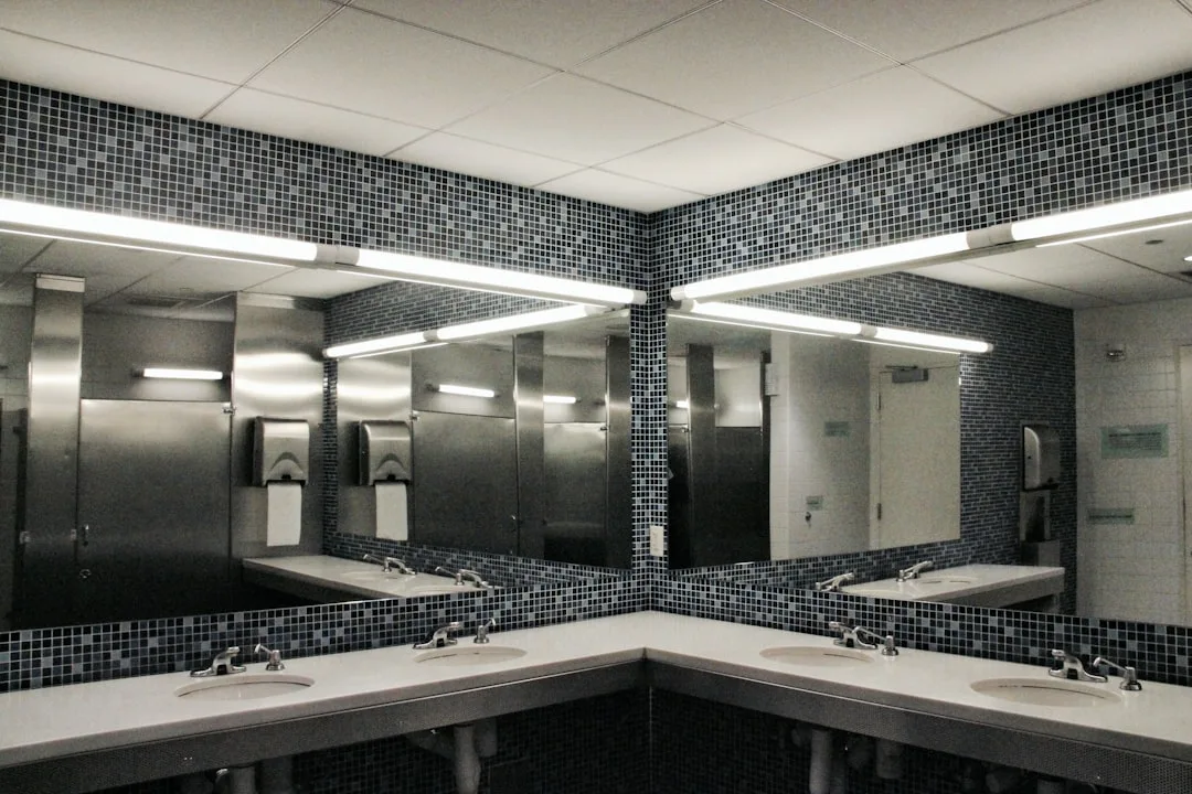 Commercial bathroom with fluorescent strip lighting mounted above large mirrors over white sinks with mosaic tile walls