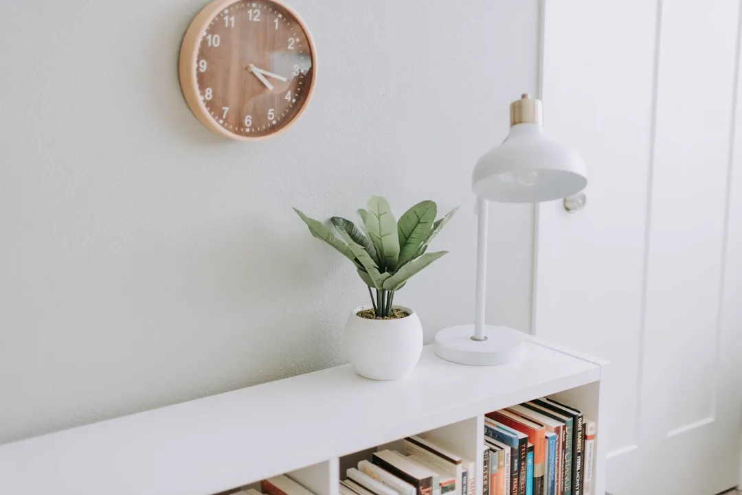 Minimalist white bookshelf with styled plant and lamp beside wall clock in a clean home office setup