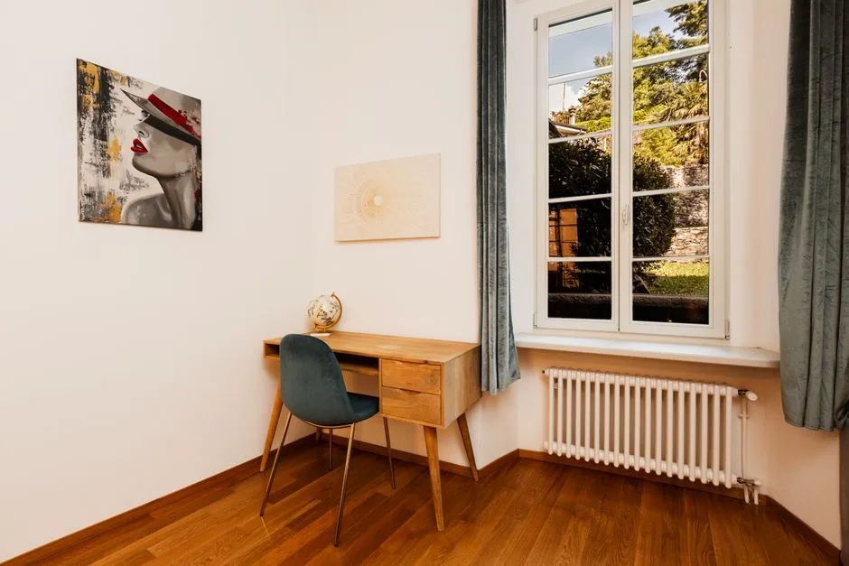 Mid-century wooden desk with teal velvet chair placed in small room corner next to window with radiator below